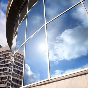 Puede incluir: Un gran edificio con una fachada de vidrio curvada que refleja un cielo azul brillante con nubes blancas. El sol brilla intensamente y se refleja en el vidrio.