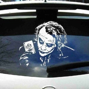 Op de afbeelding: Een witte vinyl sticker van de Joker uit de film The Dark Knight die een speelkaart met de Joker erop vasthoudt. De sticker zit op de achterruit van een auto.