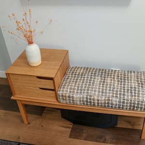 Pu&ograve; includere: Una panca in legno con un sedile imbottito e un piccolo cassetto sottostante. La panca &egrave; realizzata in legno chiaro e ha una finitura naturale. Un vaso con fiori secchi &egrave; posto sopra il cassetto.