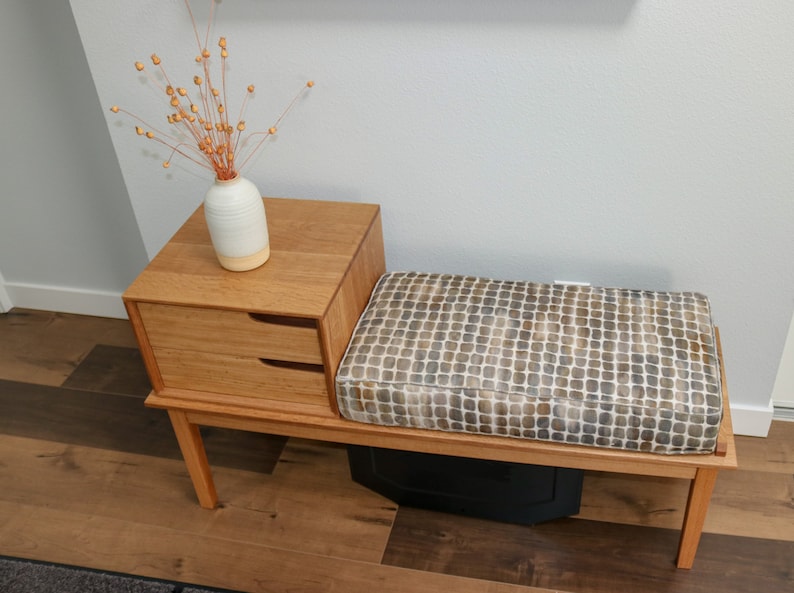 Pu&ograve; includere: Una panca in legno con un sedile imbottito e un piccolo cassetto sottostante. La panca &egrave; realizzata in legno chiaro e ha una finitura naturale. Un vaso con fiori secchi &egrave; posto sopra il cassetto.