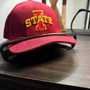 May include: A maroon baseball cap with the word "STATE" in yellow embroidery. The cap is resting on a black object, likely a pair of glasses, on a dark wooden surface. The cap has a structured, athletic design.