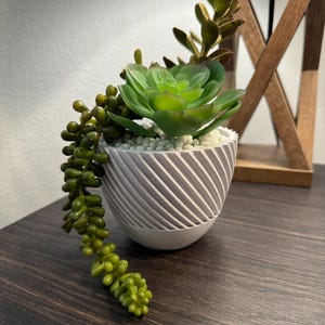 May include: A small gray planter with a striped pattern holds a succulent plant with green leaves. The plant has long trailing stems with small green leaves.