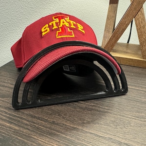 May include: A black baseball cap holder with three tiers, holding a red baseball cap with a yellow "I STATE" logo.