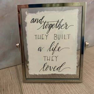 May include: A framed print with a white background and black calligraphy that reads "and together they built a life they loved".