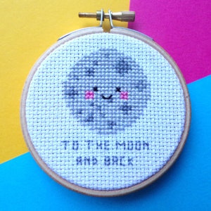 May include: A white embroidery hoop with a gray moon with a smiling face and pink cheeks. The text "To the moon and back" is embroidered below the moon.