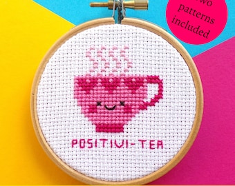 May include: A pink and white cross stitch pattern of a teacup with steam coming out of it. The teacup has a happy face and the text "POSITIVI-TEA" is stitched below it. The pattern is in a wooden embroidery hoop.