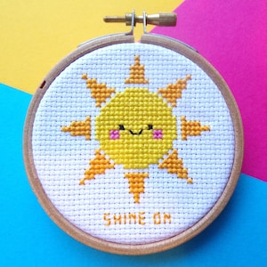 May include: A white embroidery hoop with a yellow sun with a smiling face and pink cheeks. The sun has orange rays and the text "SHINE ON" is embroidered below the sun.