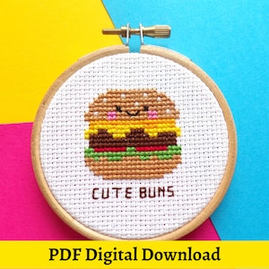 May include: A white embroidery hoop with a cross-stitch design of a smiling hamburger with the text "Cute Buns".