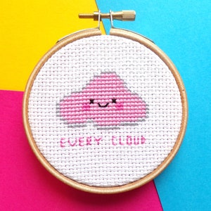 May include: A pink cloud with a smiling face embroidered on white fabric in a wooden embroidery hoop. The text "EVERY CLOUD" is stitched below the cloud.