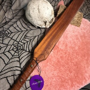 Puede incluir: Una paleta de madera marrón con un mango envuelto en cuero y tachuelas de latón. Una etiqueta morada con el texto "Raven Works" está adjunta. Una calavera y piel rosa están en el fondo.