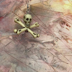 May include: A white skull and crossbones earring with a silver hook. The skull has black eye sockets and a detailed bone structure.