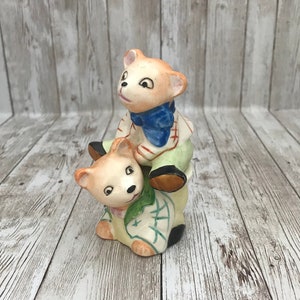 May include: A ceramic figurine of two bears, one riding on the other's back. The bear on top is wearing a green and white shirt with a blue bow tie. The bear on the bottom is wearing a green and white shirt with a green and white patterned vest.