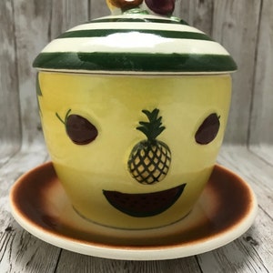 May include: A yellow ceramic sugar bowl with a lid and a matching saucer. The bowl is decorated with a smiling face, a pineapple, and a banana. The lid has a green and white striped pattern.