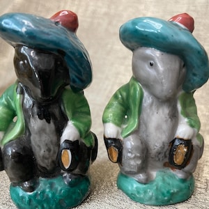 Pode incluir: Duas estatuetas de cerâmica de pequenos animais vestindo jaquetas verdes e chapéus azuis com detalhes vermelhos. As estatuetas estão sentadas em bases verdes.