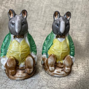 May include: Two ceramic figurines of mice wearing green jackets and yellow vests. The mice are sitting on a base and have black eyes and noses.