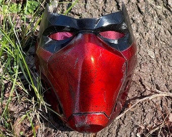 May include: A red and black plastic mask with a textured surface and a black visor. The mask has a pointed chin and a raised brow.