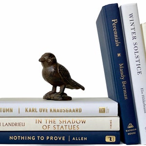 May include: A stack of books with white and blue covers. The titles of the books are visible: "The Invoice", "Winter Solstice", "Perennials", "Autumn", "In the Shadow of Statues", and "Nothing to Prove". A small brown bird figurine sits on top of the books.