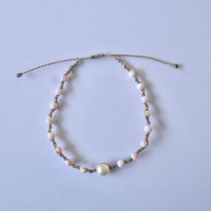 May include: A delicate bracelet made with natural materials. It features a single large pearl bead and smaller pink and white beads strung on a beige cord.