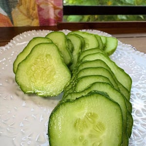 May include: Freshly sliced green cucumbers arranged on a white, decorative plate. The cucumber slices are round and show the inner seeds. The plate has a delicate, lacy pattern. The background includes a window and a glimpse of greenery.