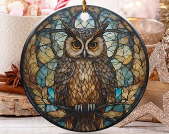 May include: A round Christmas ornament featuring a detailed owl design. The owl is depicted with brown and gold feathers, set against a stained-glass style background with blue and gold accents. The ornament hangs from a gold string.