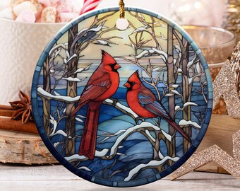 May include: A round Christmas ornament featuring two red cardinals perched on a snow-covered branch. The ornament has a stained-glass effect with blue, white, and brown tones. A gold string is attached for hanging.