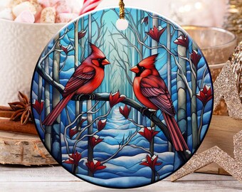 May include: A round Christmas ornament featuring two red cardinal birds perched on a branch in a winter forest scene. The ornament has a gold string for hanging. The background includes a cup of marshmallows, candy canes, and other holiday decorations.