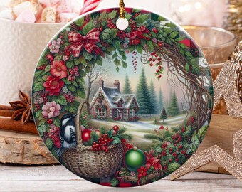 May include: A round Christmas ornament featuring a winter scene with a house, trees, and a basket of fruit. The ornament is decorated with red and green foliage and a red plaid bow. A gold string is attached for hanging.