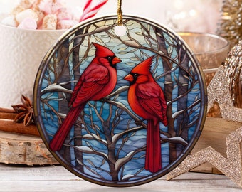 May include: A round Christmas ornament featuring two red cardinals perched on a snow-covered branch. The ornament has a gold-colored border and a gold string for hanging. The background shows a winter scene with bare trees.