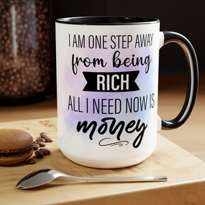 May include: White ceramic mug with a black interior and handle. The mug features the text "I AM ONE STEP AWAY from being RICH ALL I NEED NOW IS money" in black script. A watercolor-style purple and blue design is behind the text.