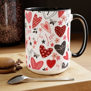 May include: A large white ceramic mug with a black interior and handle. The mug is decorated with a pattern of red and pink hearts, stars, flowers, and the word "Love". A spoon and a macaron are in the foreground.