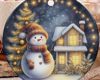 May include: A round Christmas ornament featuring a snowman, a house, and a Christmas tree under a starry night sky. The snowman wears a brown hat and scarf. The ornament is hanging with a gold string. Candy canes and a cup of marshmallows are in the background.