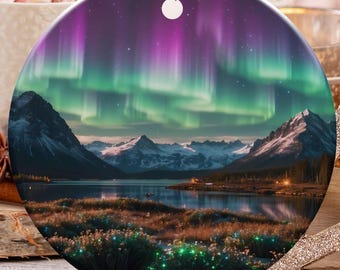 May include: A round Christmas ornament with a scenic landscape of mountains, a lake, and the aurora borealis. The ornament is hanging with a gold string. Other holiday decorations are in the background.