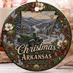 May include: A round ceramic Christmas ornament featuring a green pickup truck carrying a pine tree, a map of Arkansas, and the text "Christmas in Arkansas" and "Natural State Holiday Keepsake" surrounded by a wreath of pinecones, berries, and flowers.