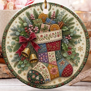 Op de afbeelding: Een ronde kersthanger van keramiek met een kleurrijk patchwork-kerstsokontwerp. De sok is versierd met bloemmotieven, een rode strik en twee gouden belletjes, omringd door dennentakken, bessen en kleine cadeautjes.