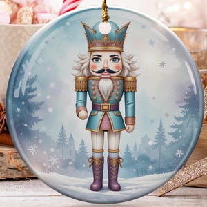 Puede incluir: Adorno navideño redondo con una ilustración en acuarela de un soldado cascanueces con uniforme azul y rosa, sobre un fondo de bosque nevado. El adorno cuelga de un hilo dorado. Otras decoraciones navideñas en el fondo.