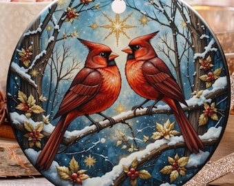 Peut inclure: Ornement de Noël rond avec deux cardinaux rouges perchés sur une branche enneigée. Le fond représente une scène de forêt hivernale avec un ciel bleu et de la neige qui tombe. L'ornement est suspendu par une ficelle dorée.