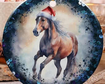 May include: A round Christmas ornament featuring a watercolor painting of a brown horse wearing a Santa hat. The ornament has a blue sequined border and a gold string for hanging. The background includes candy canes, a mug of marshmallows, and other holiday decorations.