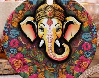May include: A round Christmas ornament featuring a colorful illustration of the Hindu deity Ganesha. The ornament has a gold string for hanging and is decorated with floral patterns. The background includes holiday decorations and a cup of hot cocoa.