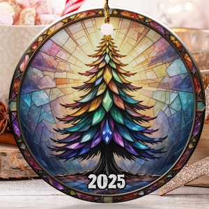 Puede incluir: Adorno navideño redondo con un diseño de vidriera colorido que representa un árbol de Navidad. El adorno tiene una cuerda dorada para colgar y el año "2025" en la parte inferior. Bastones de caramelo y otras decoraciones navideñas están en el fondo.