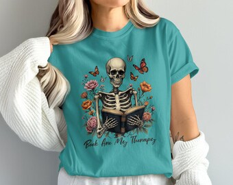 May include: Teal t-shirt with a skeleton reading a book, encircled by flowers and butterflies. The skeleton is the main design, with the text "Book Are My Therapy" beneath. The shirt is a comfortable, casual style, suitable for everyday wear.