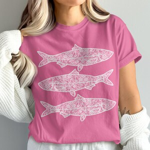 May include: A pink t-shirt featuring three white fish designs. Each fish is filled with a detailed, floral pattern. The shirt has short sleeves and a crew neck. The overall design is simple and eye-catching.