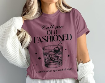 Peut inclure: T-shirt mauve avec l'inscription "Call me OLD FASHIONED" et un dessin d'un cocktail Old Fashioned avec une cerise, une tranche d'orange, des glaçons et des étoiles. La phrase "classic never goes out of style" est en dessous.