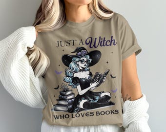May include: Tan t-shirt with a graphic of a witch reading a book, wearing a black hat and dress, and glasses. The text reads "JUST A Witch WHO LOVES BOOKS". The witch has blue hair and is surrounded by bats and stars.