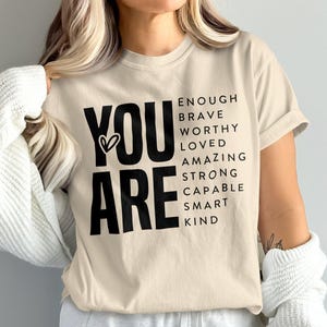 May include: A beige t-shirt with the words "YOU ARE" in large black letters, with a heart symbol between the words. Below, smaller text lists positive affirmations: ENOUGH, BRAVE, WORTHY, LOVED, AMAZING, STRONG, CAPABLE, SMART, KIND.