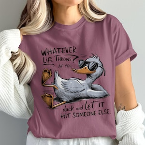 Puede incluir: Camiseta color malva con un dibujo de un pato de dibujos animados con gafas de sol y el texto "Whatever life throws at you, duck and let it hit someone else." El pato es gris y blanco con pico y patas amarillas.
