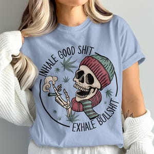 May include: Pale blue t-shirt with a skull wearing a red and green striped scarf and beanie, smoking a cigarette. The shirt has the text "INHALE GOOD SHIT EXHALE BULLSHIT" in a circular design with cannabis leaves.