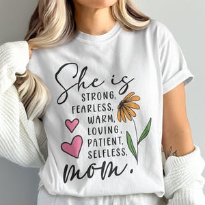 Może przedstawiać: Biała koszulka z napisem "She is STRONG, FEARLESS, WARM, LOVING, PATIENT, SELFLESS, mom." czarnym pismem. Projekt zawiera dwa różowe serca oraz żółto-pomarańczowy kwiat z zielonymi liśćmi.