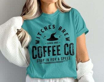 Puede incluir: Camiseta de color turquesa con un gráfico negro que dice "Witches Brew Coffee Co" con una ilustración de un sombrero de bruja. El texto incluye "Since 1962" y "Stop in for a Spell". La camiseta tiene mangas cortas y cuello redondo.