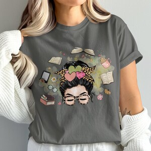 May include: Grey t-shirt with a graphic of a woman with glasses and a messy bun, surrounded by books, a coffee cup, and flowers. The woman's hair is styled with a leopard print headband and heart-shaped accents. A cute design for book lovers.