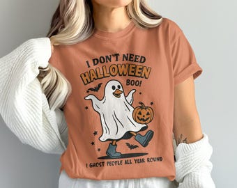 以下が含まれることがあります： ハロウィーンをテーマにしたグラフィックのオレンジ色のTシャツ。デザインは、カボチャとブーツを持った漫画の幽霊が特徴で、「I DON'T NEED HALLOWEEN BOO! I GHOST PEOPLE ALL YEAR ROUND.」というテキストが書かれています。